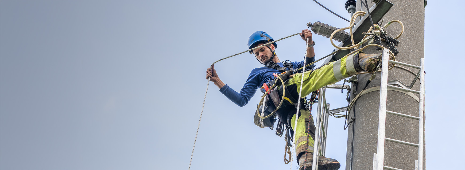 Netzelektiker hängt an Strommast und schaut in die Tiefe.